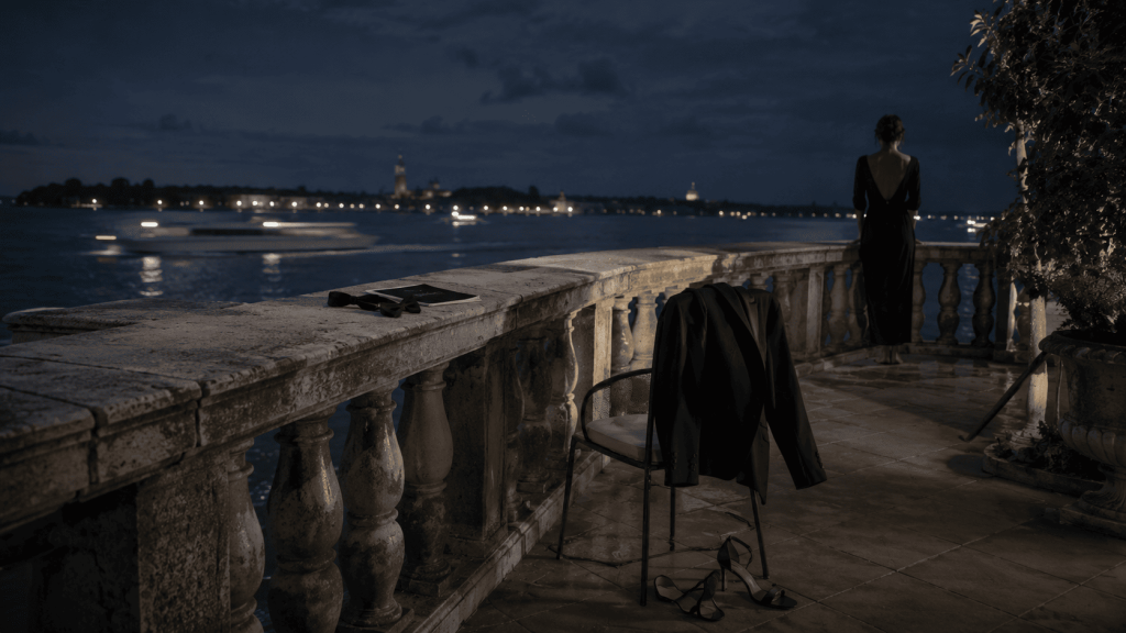 Terrazza sul Lido di Venezia dopo una première, con abito da smoking, scarpe da sera e una donna affacciata sulla laguna illuminata di notte.