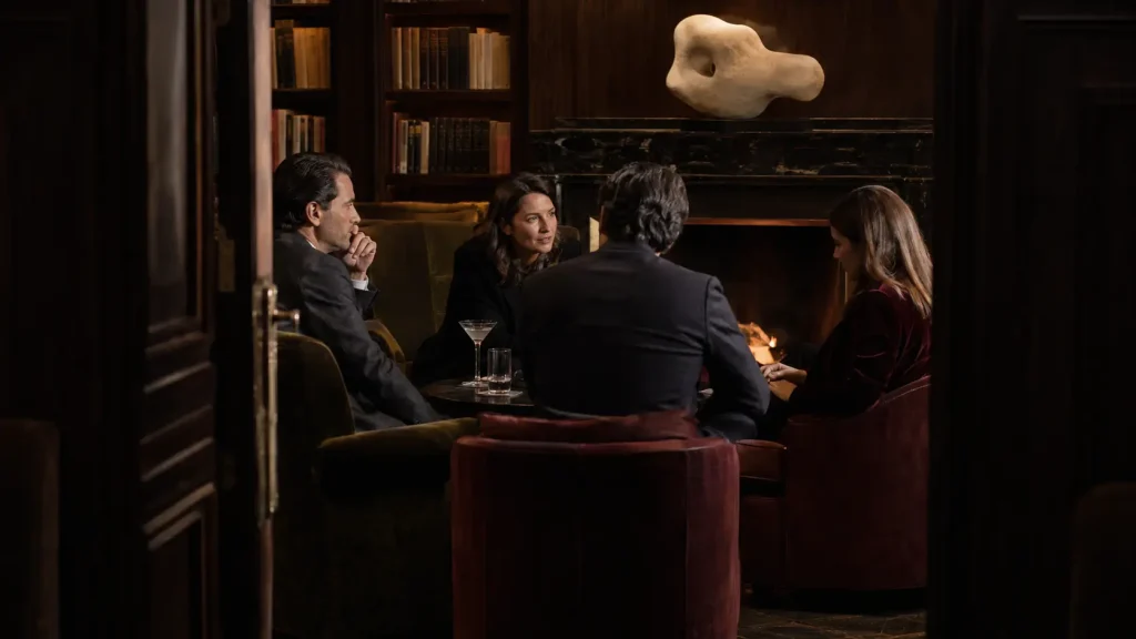 Conversazione riservata tra quattro membri in una biblioteca privata, con camino acceso, arredi in velluto, libri d’arte e atmosfera elegante.