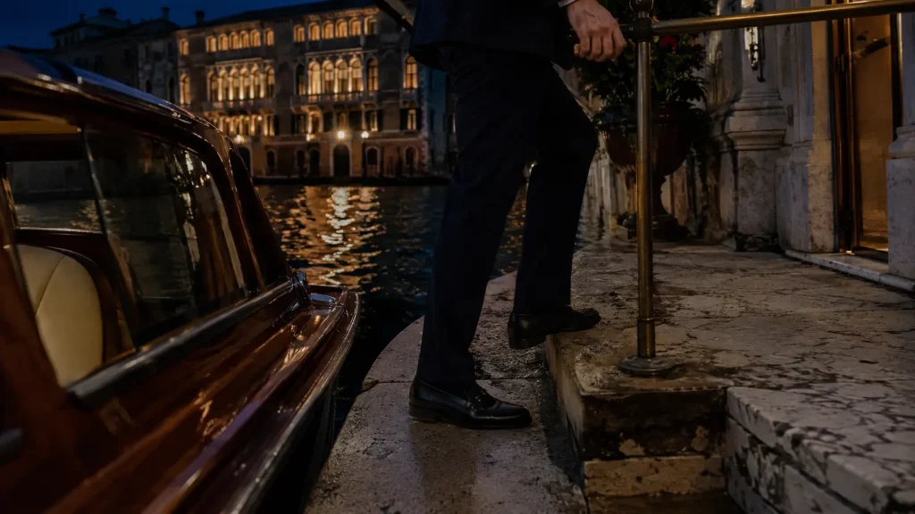 Arrivo notturno in taxi acqueo a un palazzo veneziano sul Canal Grande, con ospite elegante che scende sulla riva tra riflessi dorati sull’acqua.