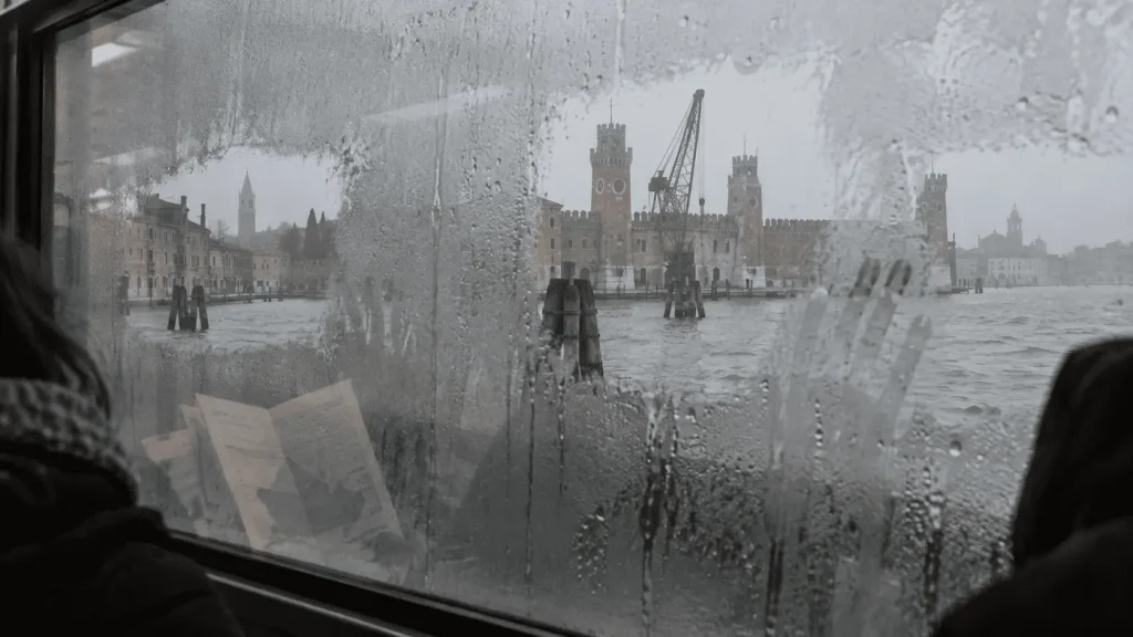 Vista autunnale di Venezia attraverso il finestrino appannato di un vaporetto, con scorci dell’Arsenale e della laguna tra condensa e riflessi.
