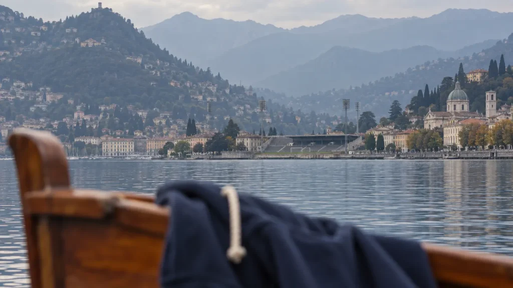 Stadio storico di Como tra architetture sul lago e montagne, con una felpa blu sfocata appesa a una barca in primo piano.