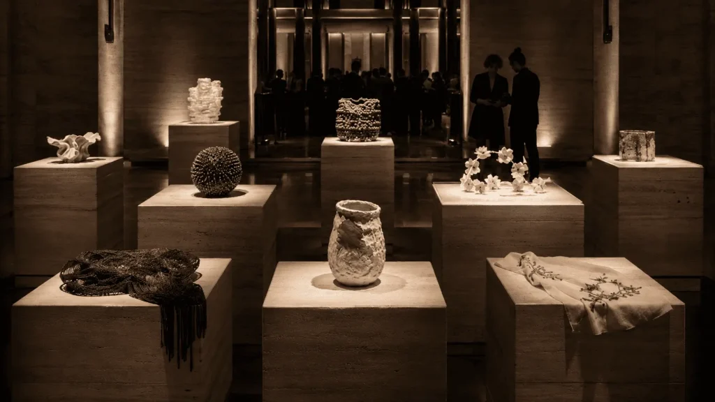 Anteprima privata di artigianato italiano contemporaneo in un elegante salone romano, con opere esposte su piedistalli in travertino e ospiti sullo sfondo.