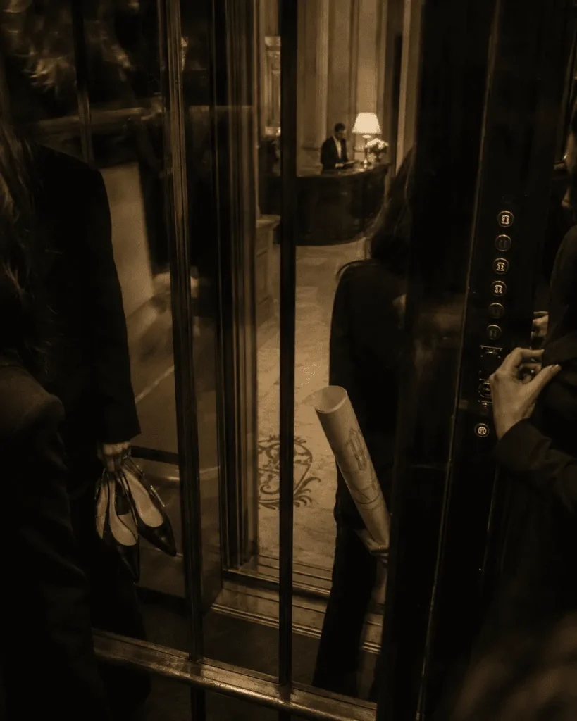 Rientro notturno durante la Milano Fashion Week, con ospiti eleganti riflessi in un ascensore d’epoca e concierge visibile nella hall.