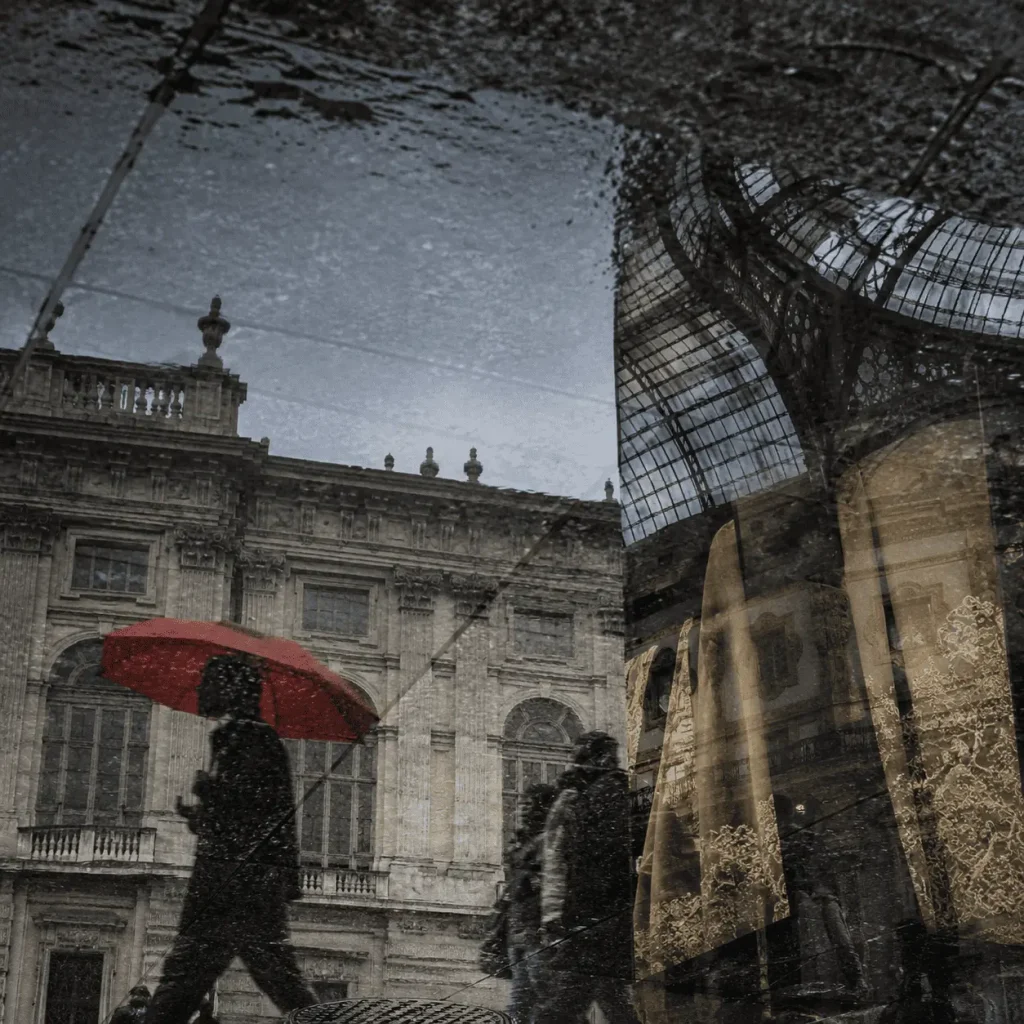 Riflessi urbani nel centro di Milano dopo la pioggia, con Palazzo Reale, la Galleria, passanti sfocati e un ombrello rosso come unico accento cromatico.