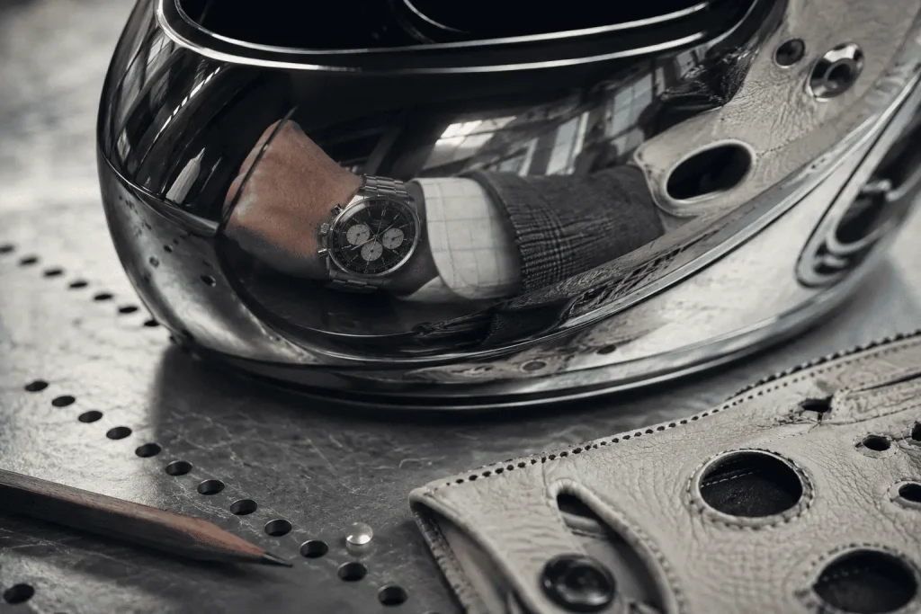 Cronografo in acciaio riflesso nella visiera di un casco da corsa, con guanto in pelle, metallo perforato e dettagli tecnici sul banco di lavoro.
