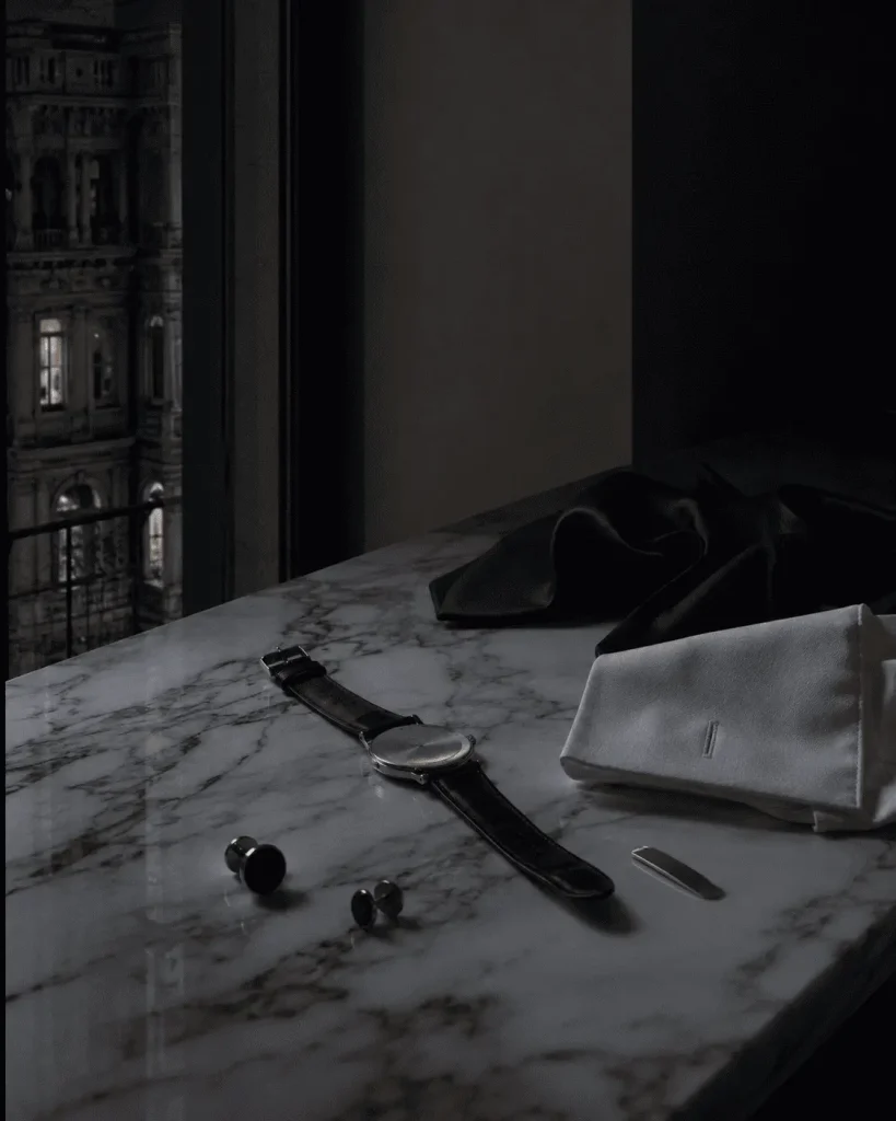 Accessori da sera su una console in marmo a Milano, con orologio, gemelli in onice, cravatta in seta e polsino bianco dopo un evento formale.